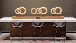 A modern hotel lobby with a reception desk, three wireframe chairs, a pendant light, and a wall-mounted display of rings. - Courtyard by Marriott New York Queens/Fresh Meadows (Fresh Meadows)