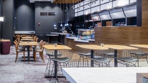 A modern cafe with wooden tables and chairs, a bar area with a menu display, and a seating area with a patterned carpet. - Courtyard by Marriott New York Queens/Fresh Meadows (Fresh Meadows)