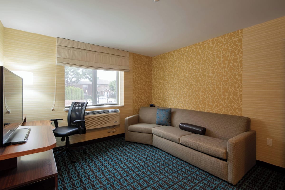 A hotel room with a wooden desk, a black office chair, a beige sofa, and a patterned carpet.