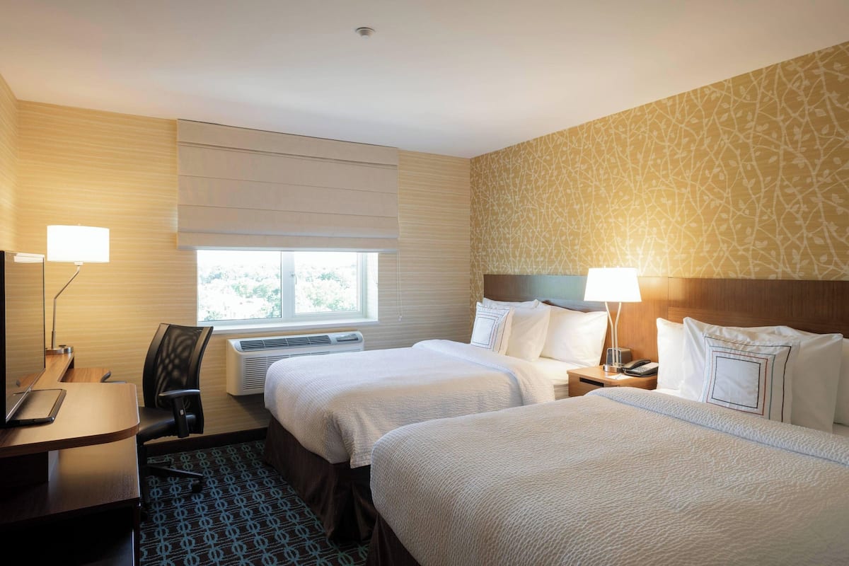 A hotel room with two beds, a desk, a chair, a lamp, and a window.