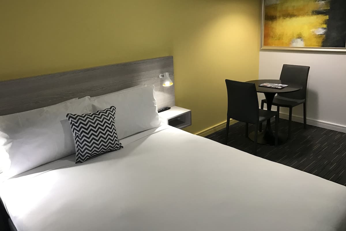 Ein Hotelzimmer mit Bett, Nachttisch mit Lampe, Tisch mit Stühlen und einem Bild an der Wand.