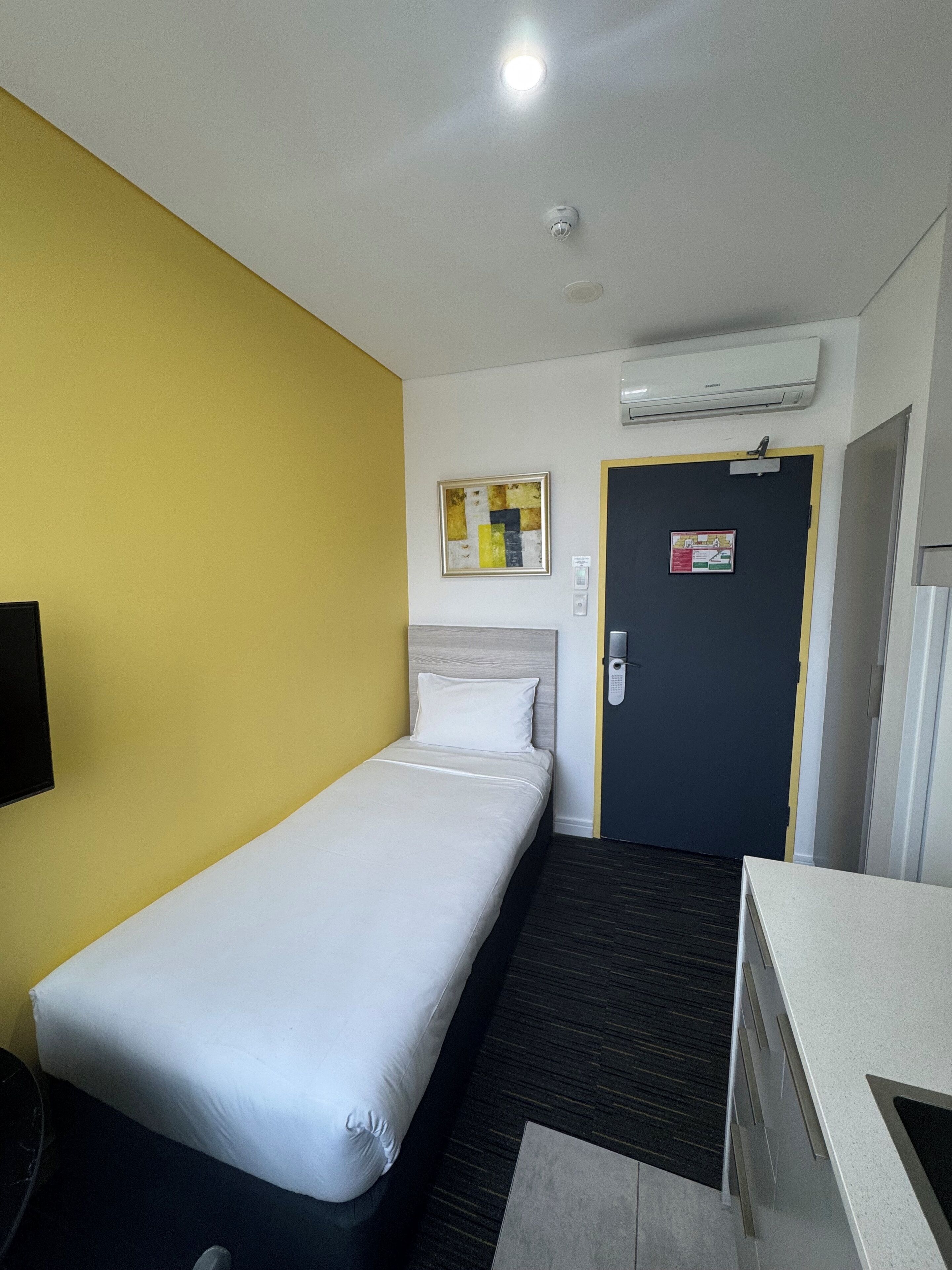 A hotel room with a single bed, a desk, a chair, a door with a fire exit sign, and an air conditioning unit on the wall.