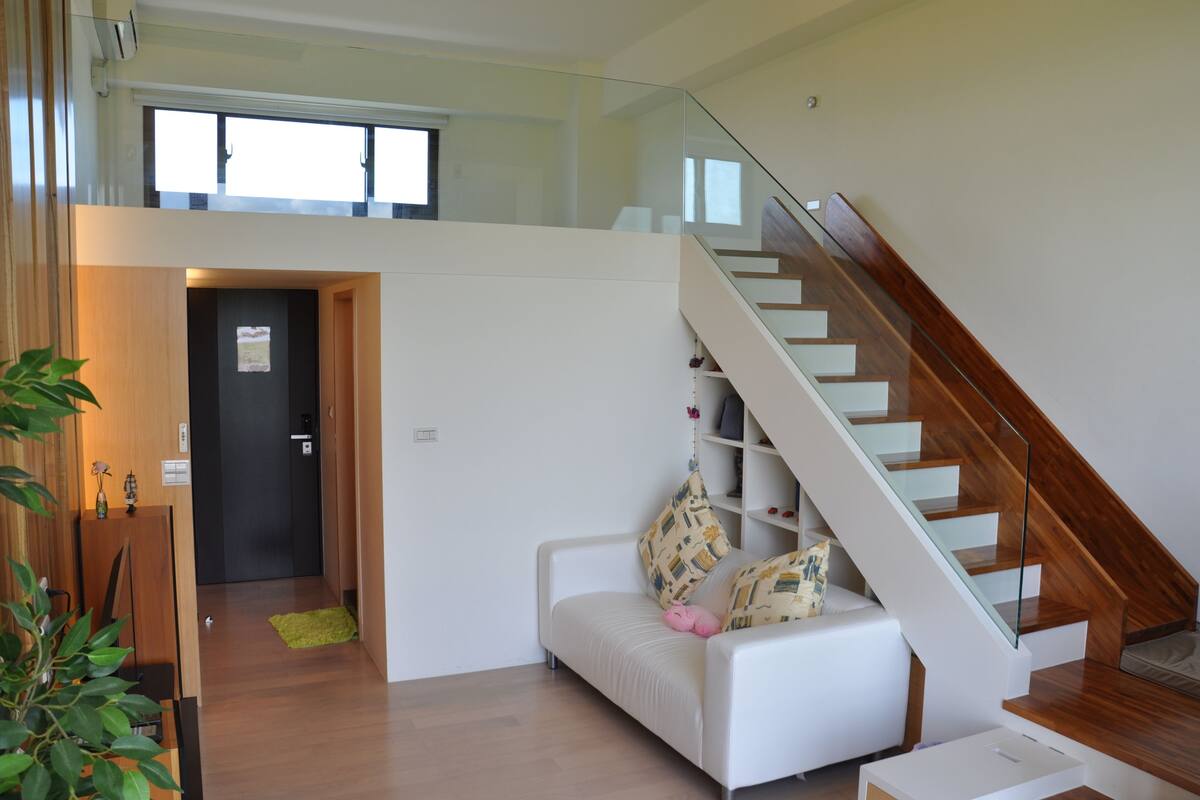 Uma sala de estar moderna com um sofá branco, uma escadaria de vidro e piso de madeira.