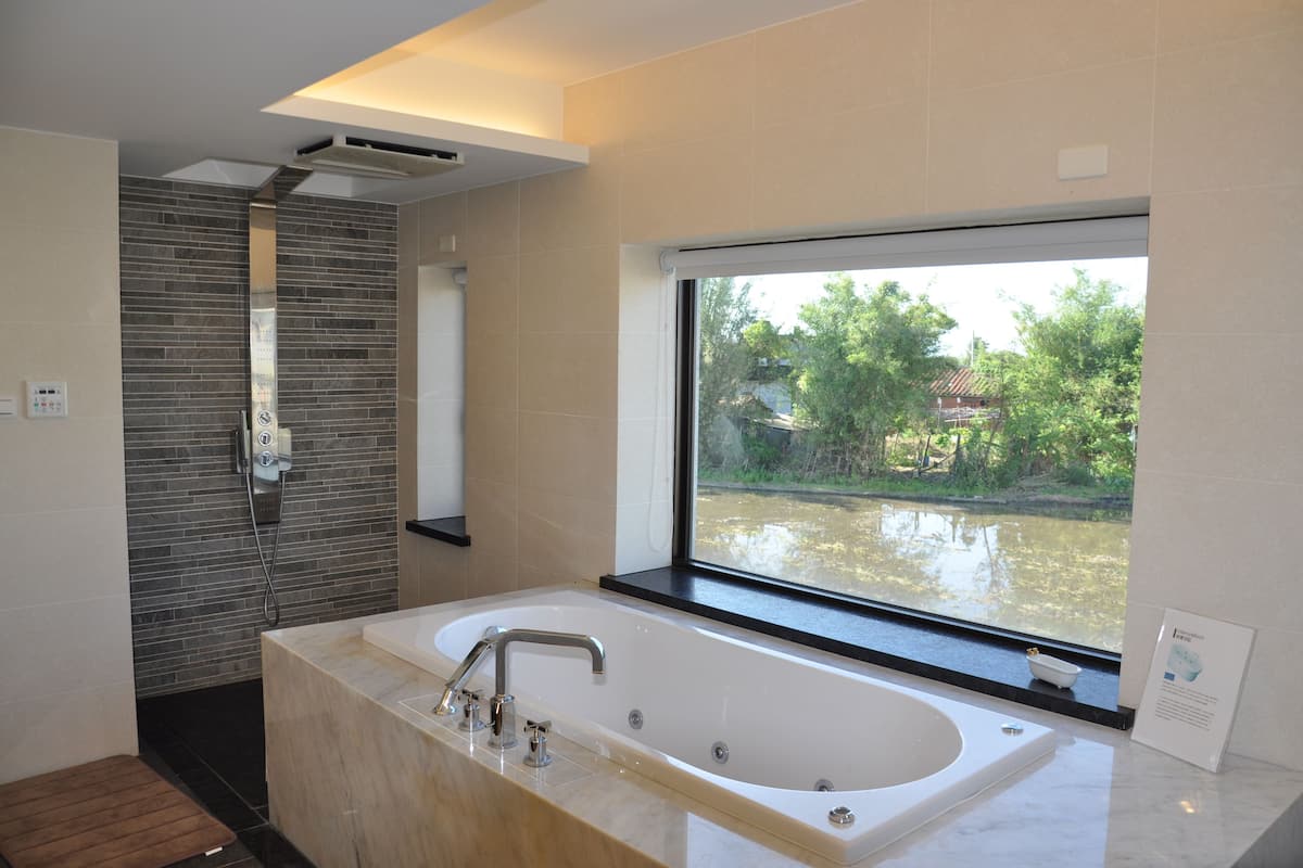 Uma casa de banho moderna com uma banheira grande, zona de duche e janela com vista para a água e vegetação.