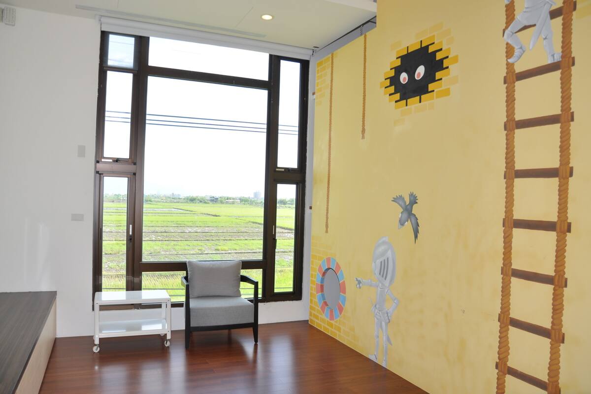 Quarto com janelas amplas, uma cadeira branca, uma pequena mesa branca e um mural colorido na parede.