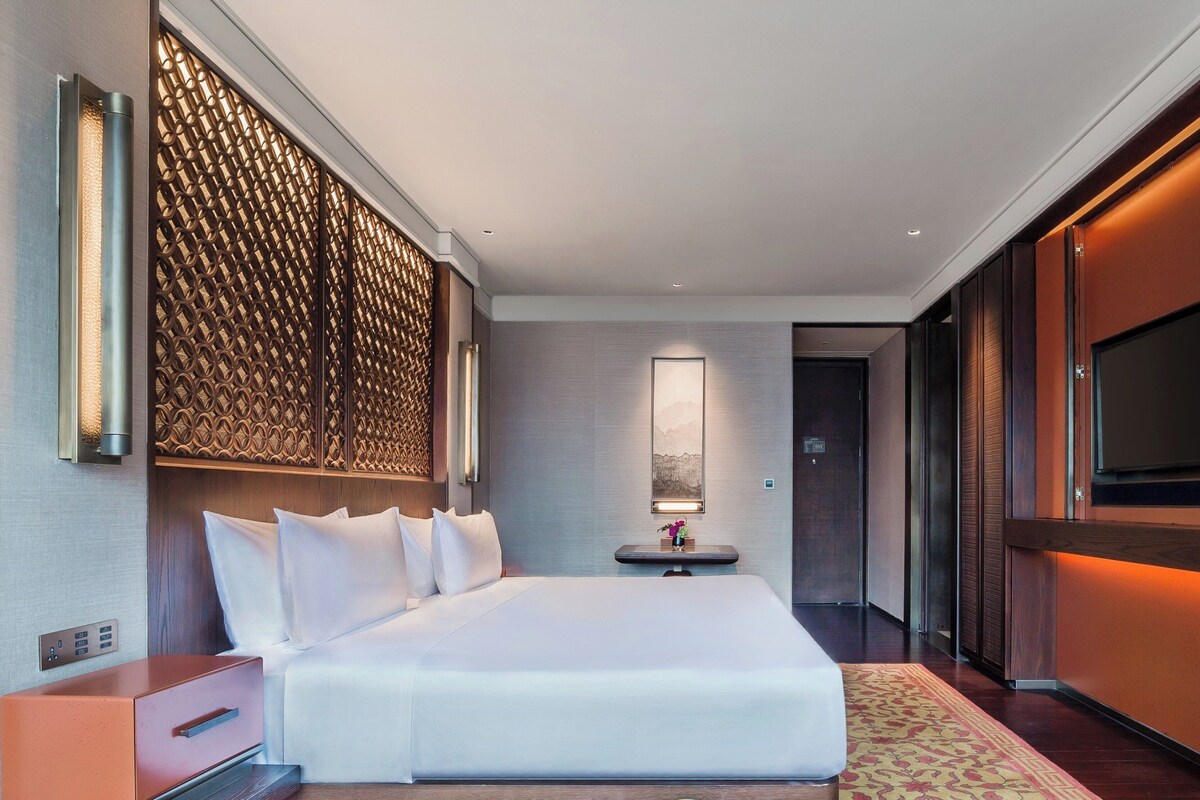 A modern hotel room with a large bed, a bedside table, a TV, and a decorative wall panel.