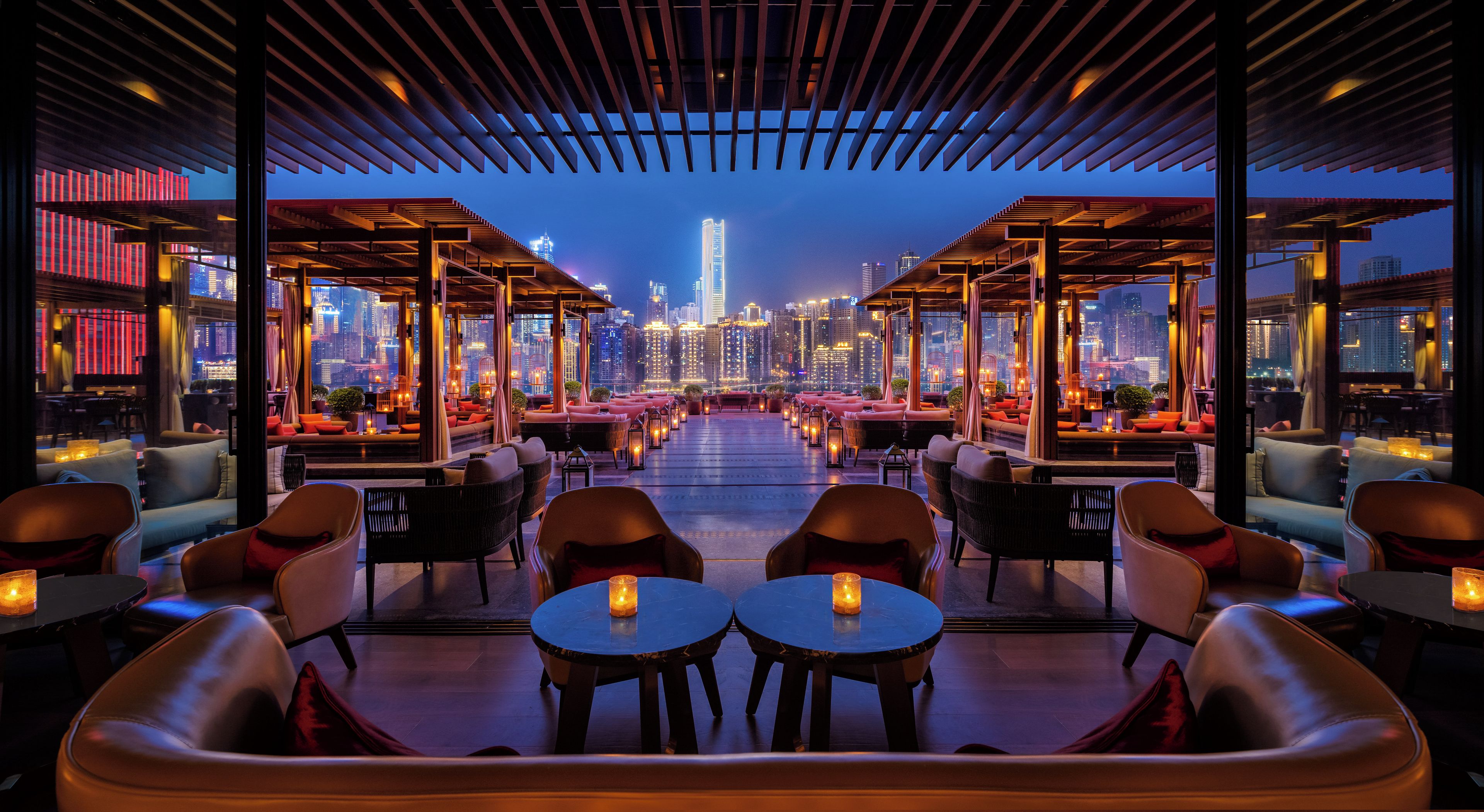 A rooftop lounge with a city view, tables, chairs, and ambient lighting.