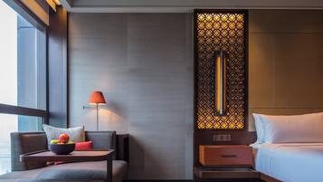 A modern hotel room with a sofa, a bedside table with fruit, a lamp, a bed, and a decorative wall panel.