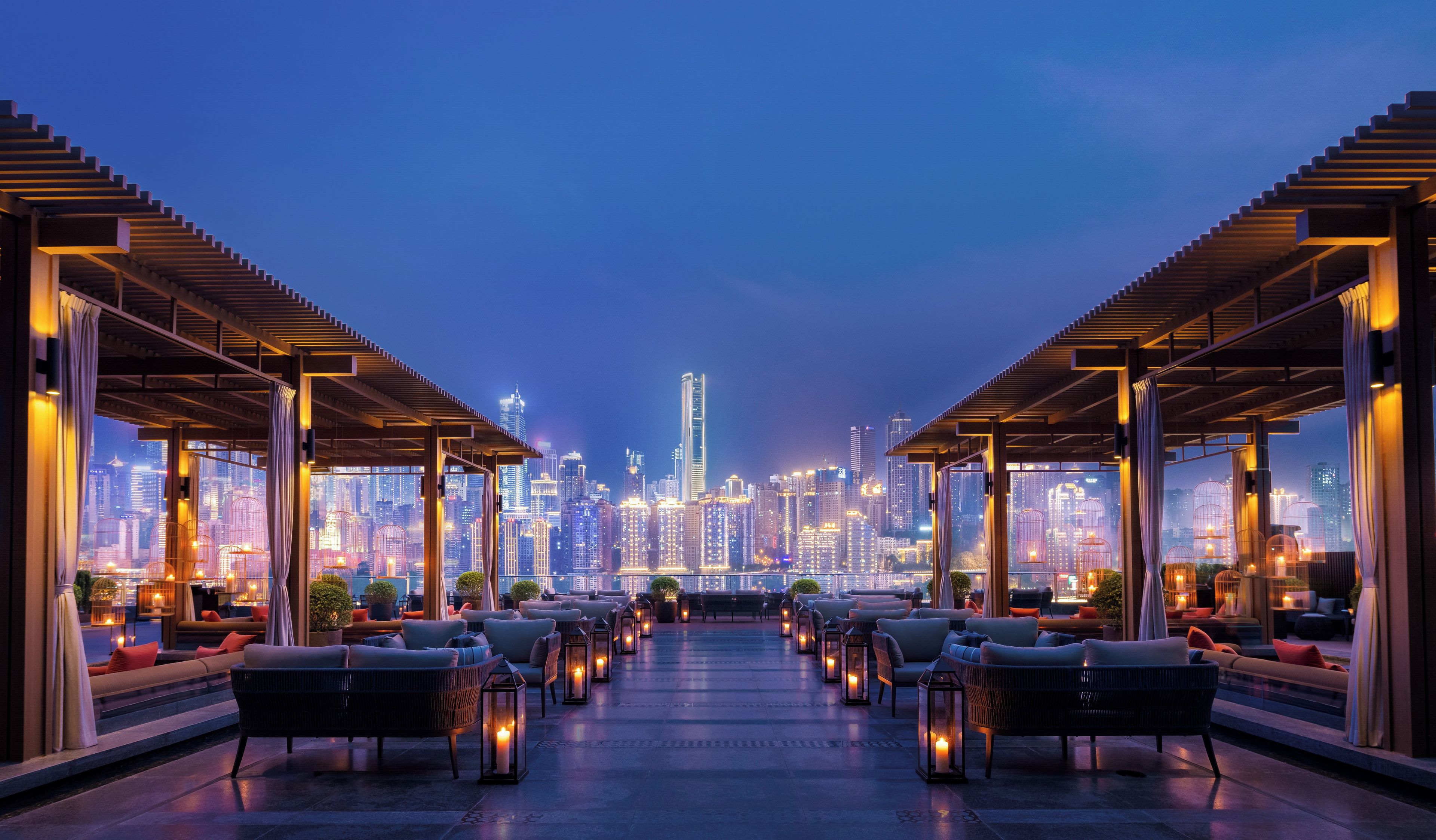 A rooftop lounge with seating and tables, overlooking a city skyline at dusk.