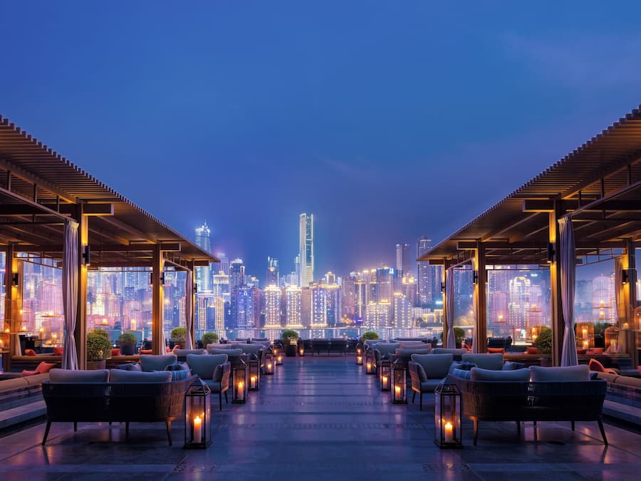 A rooftop lounge with seating and tables, overlooking a city skyline at dusk.