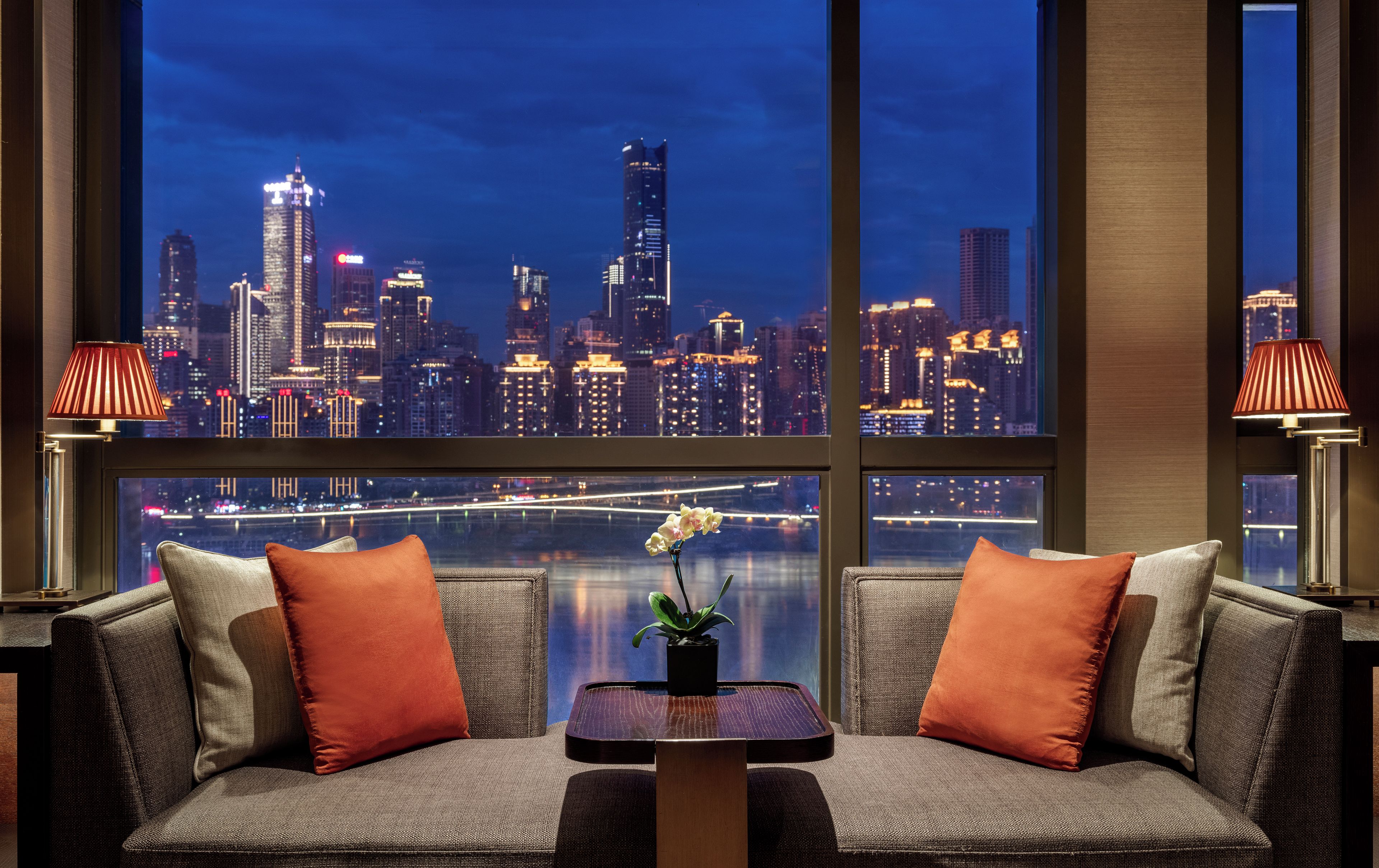 A modern living room with a sofa, two lamps, and a view of a city skyline at night.