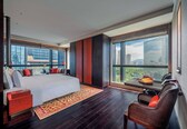 A modern hotel room with a large bed, a seating area with a table, and a view of the cityscape through large windows.