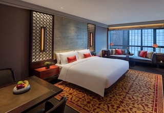 A hotel room with a large bed, a seating area with sofas, a dining table with fruit, and a view of the city through large windows.
