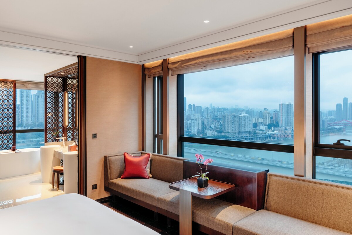 A modern hotel room with a large window offering a city view, a sofa with a red pillow, and a small table with a vase of flowers.