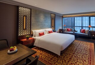 A modern hotel room with a large bed, a dining table with fruit, a sofa, and a view of the city.