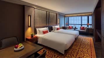 A hotel room with two beds, a dining table with fruit, a decorative wall, and a large window with a view.