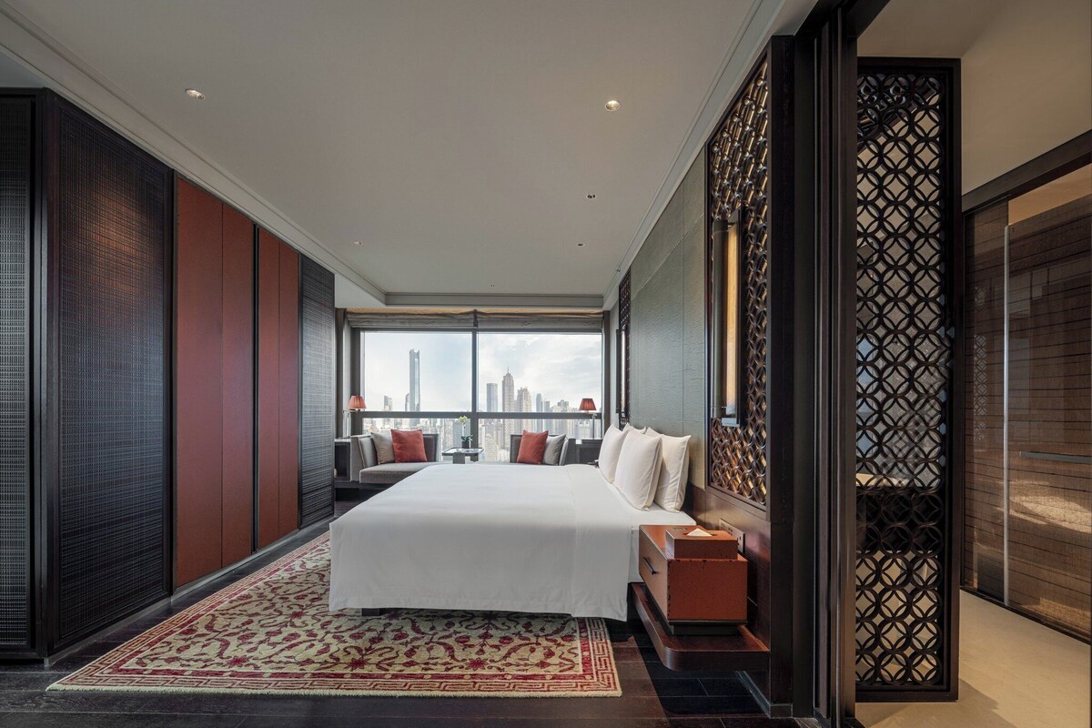 A modern hotel room with a large bed, a city view, and a traditional decorative screen.