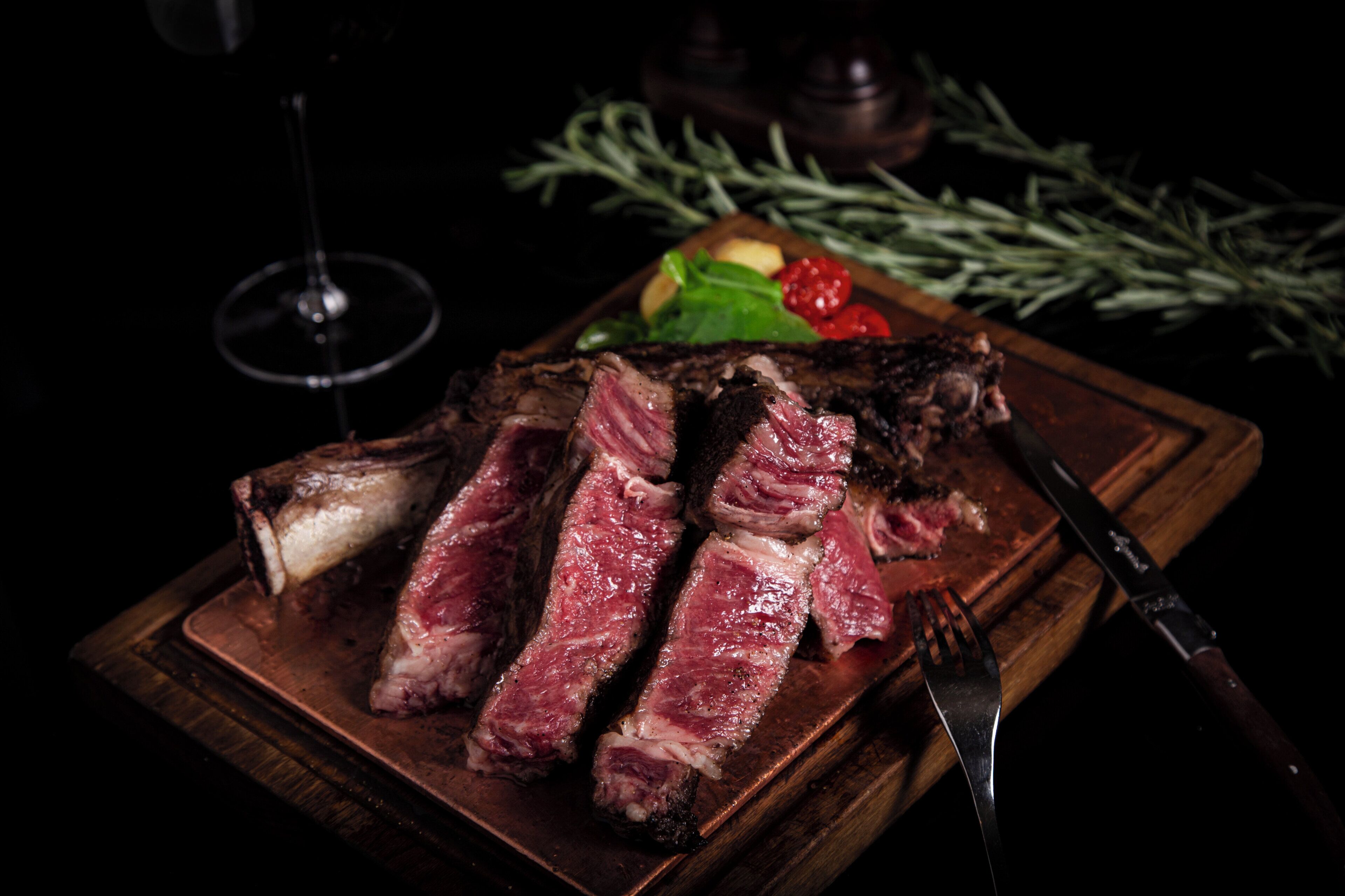 A steak with a fork and knife on a wooden board, accompanied by a glass of wine and some vegetables.