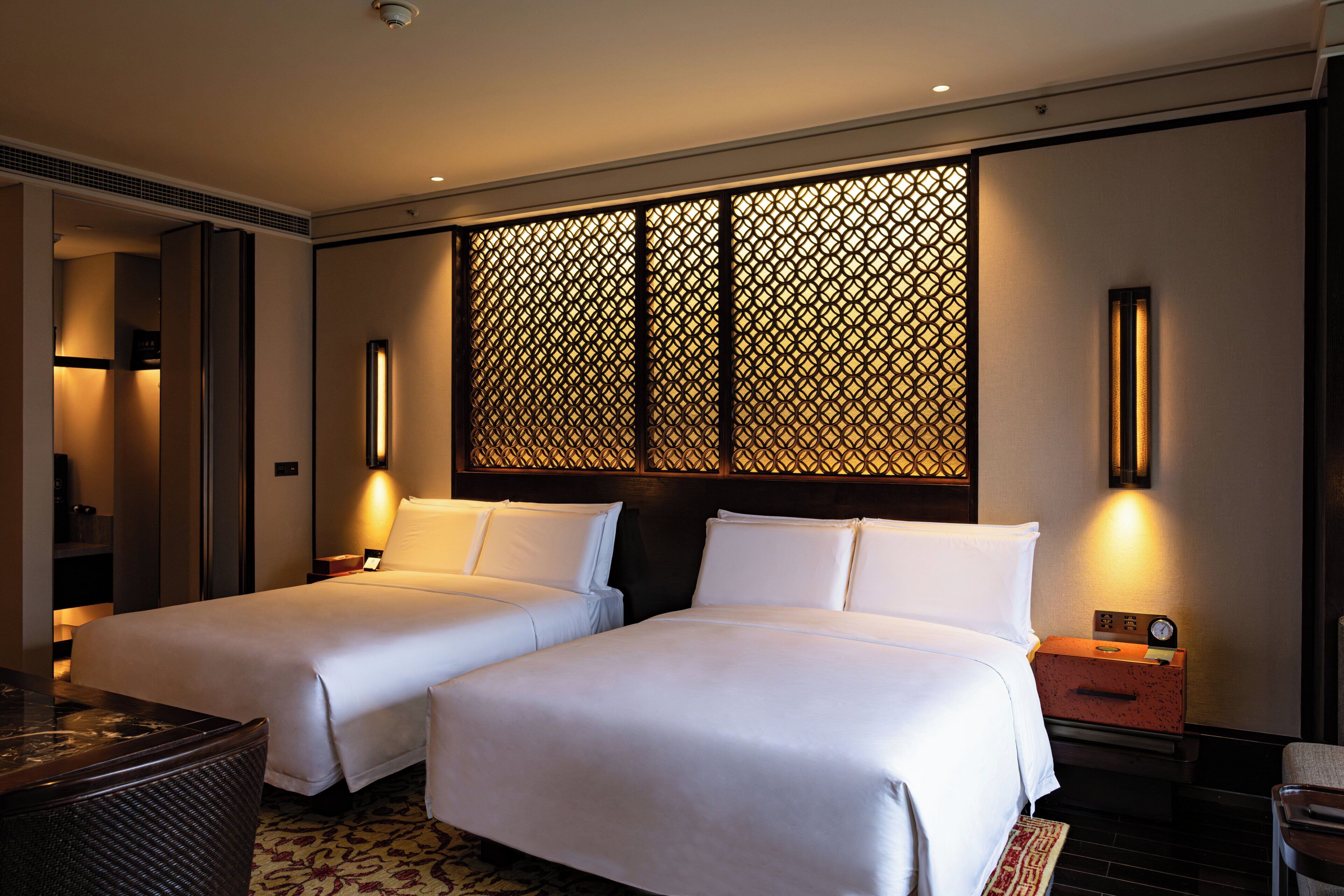 A hotel room with two beds, a nightstand, a lamp, a wall-mounted clock, and a decorative screen.