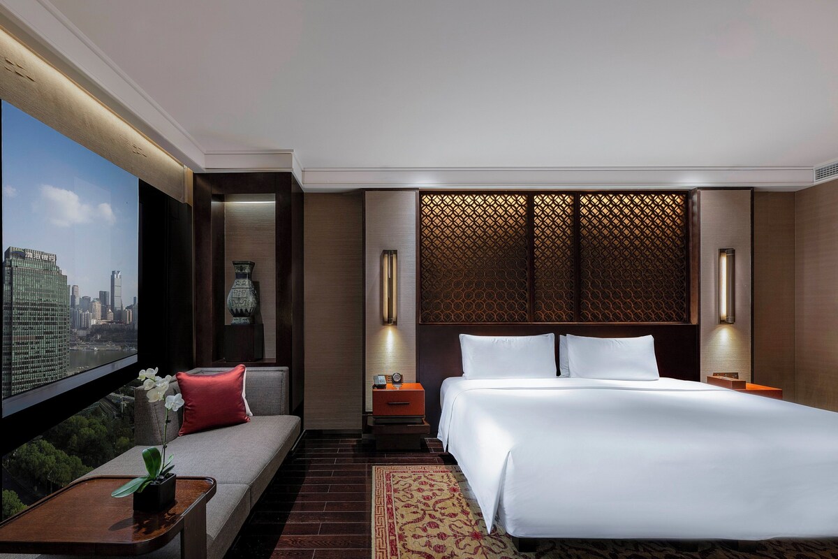 A modern hotel room with a large bed, a sofa, a TV with a cityscape view, and a decorative wall panel.