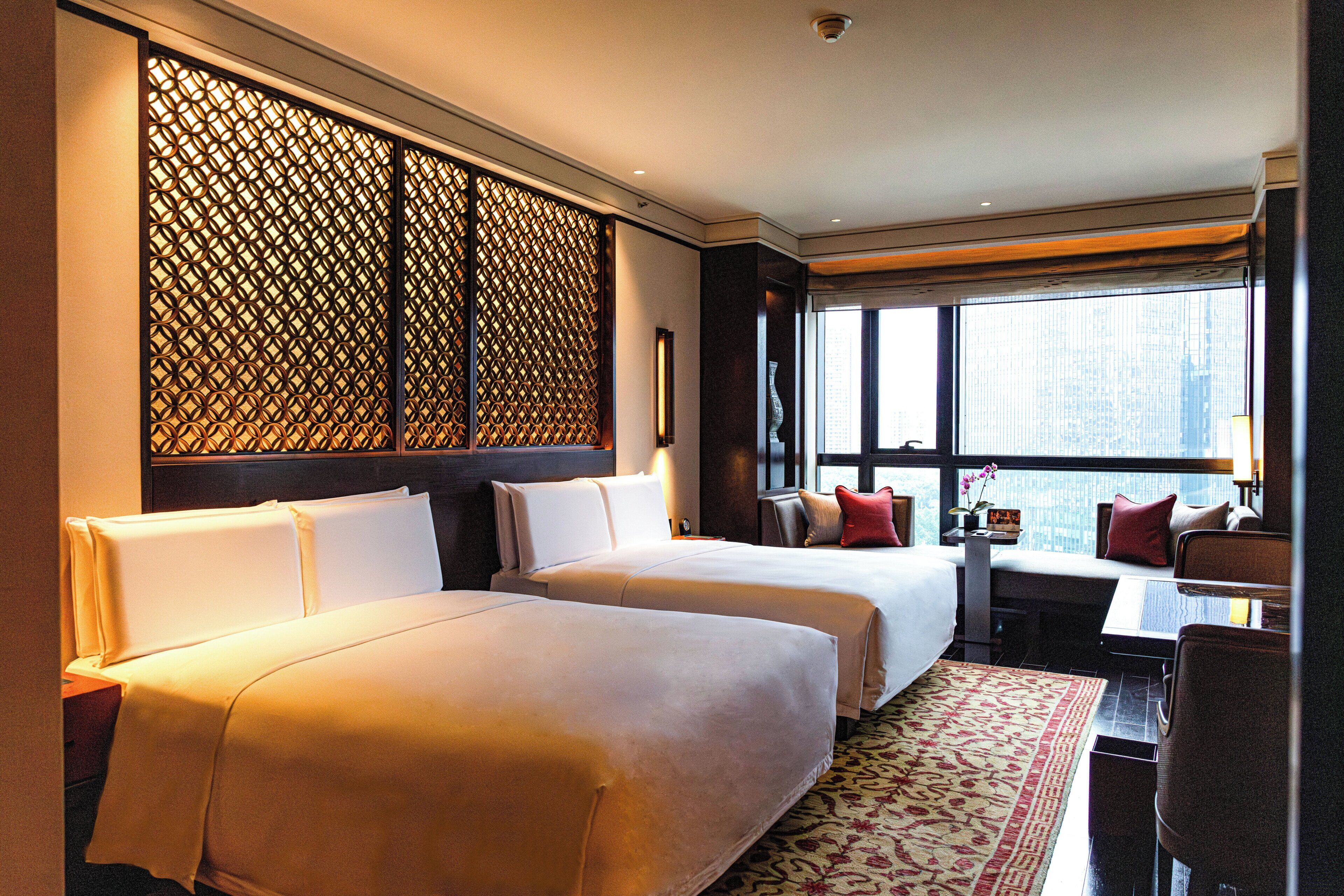 A hotel room with two beds, a desk, a chair, a window with a view, and a decorative screen.