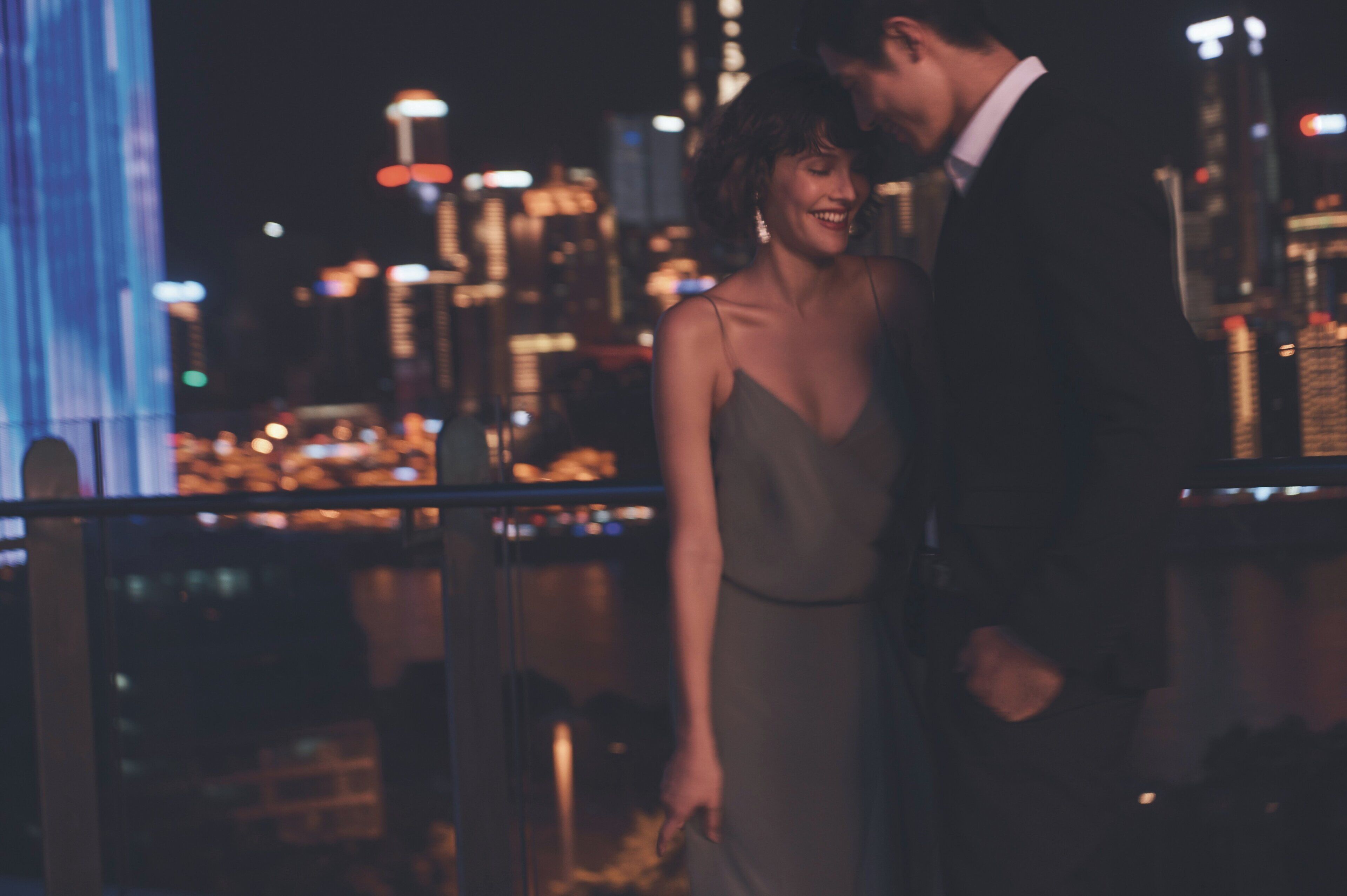 A couple standing on a balcony with a cityscape in the background.