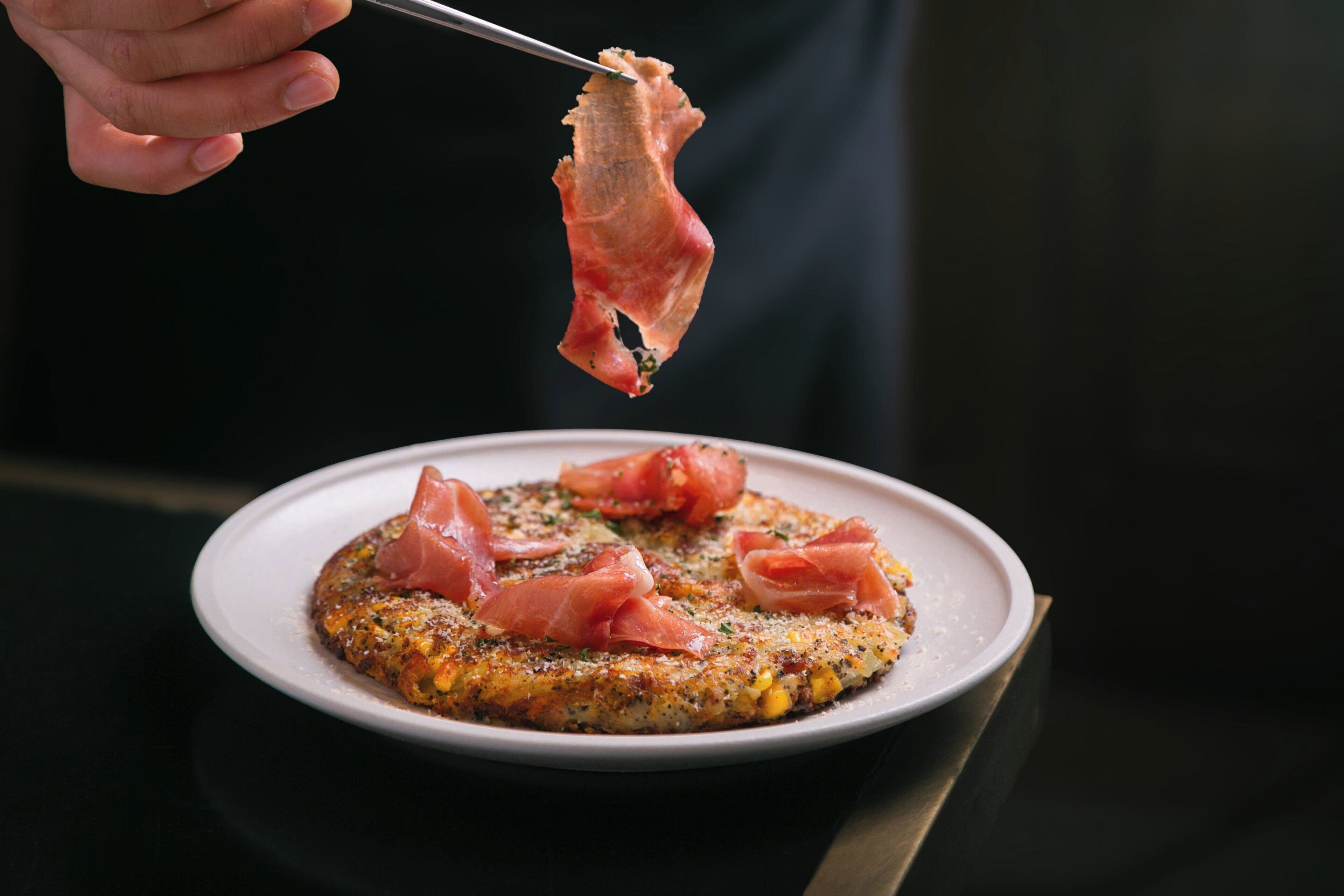 A plate with a food item topped with slices of cured meat, held by a pair of tongs.