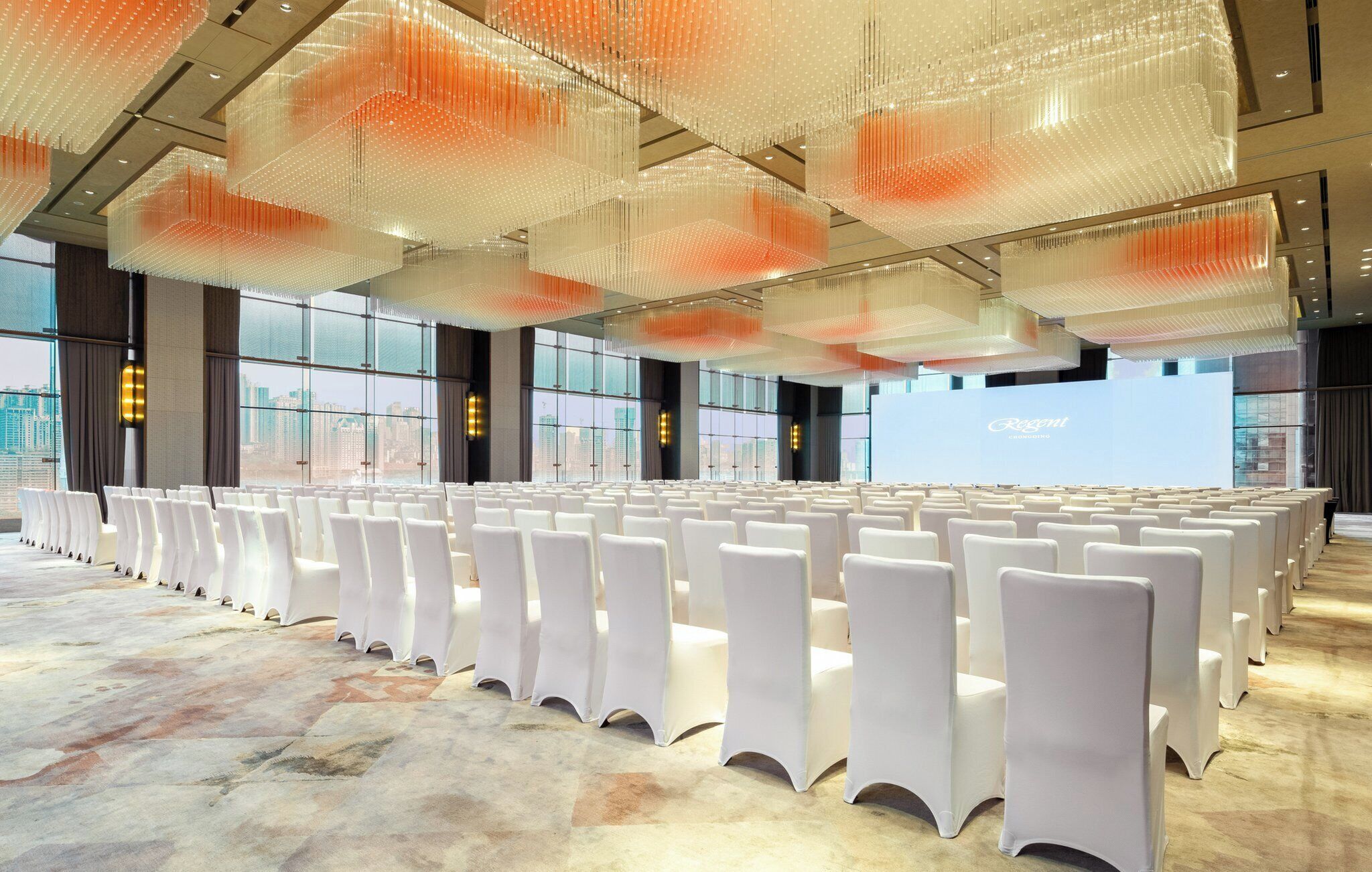 A large conference room with rows of white chairs, a large screen, and a city view.