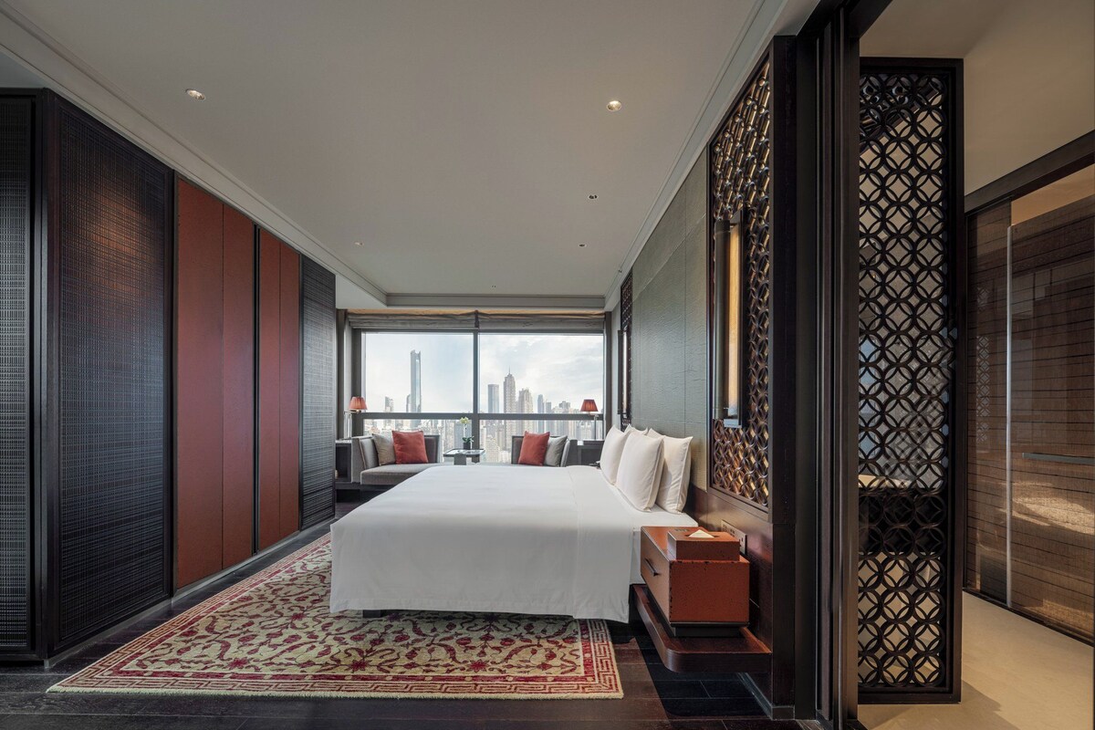 A modern hotel room with a large bed, a seating area, and a view of the city skyline.