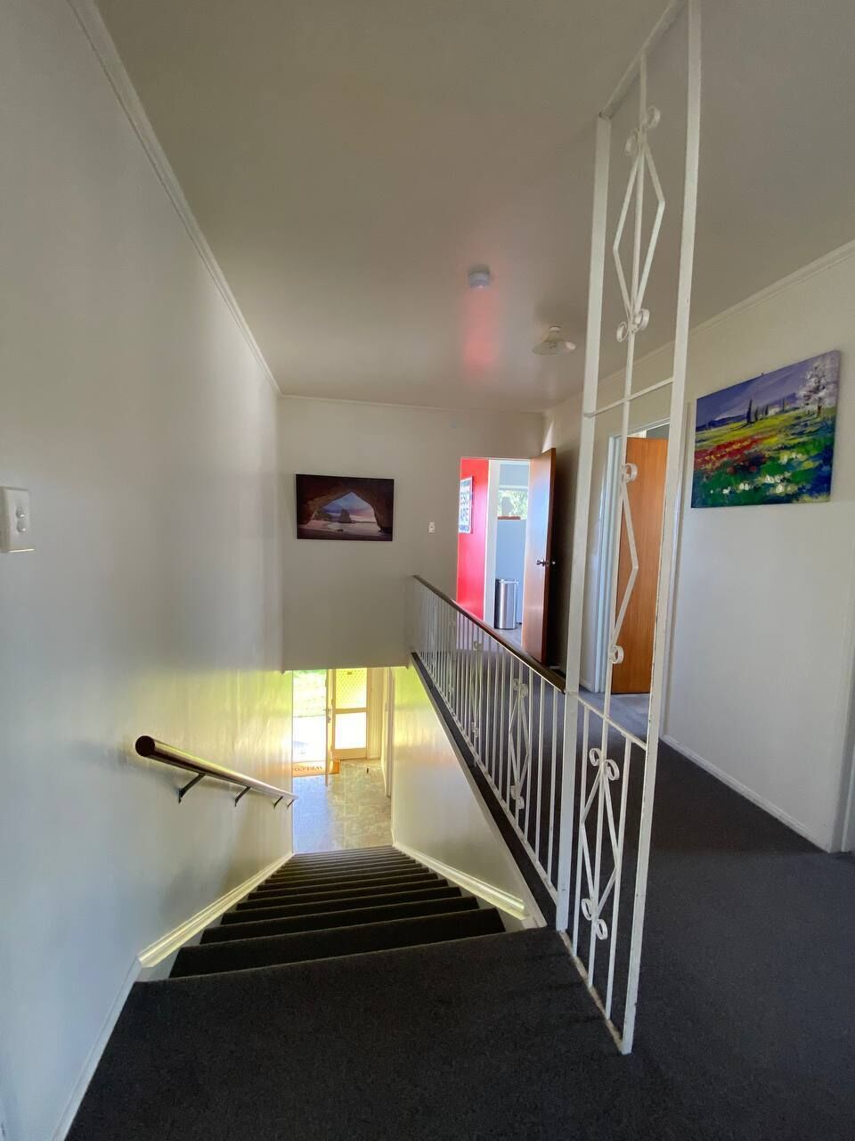 A staircase with a handrail leading to a landing, a painting on the wall, and a door with a red frame.