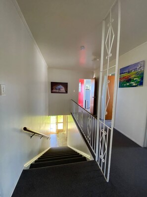 A staircase with a handrail leading to a landing, a painting on the wall, and a door with a red frame. - Waitangi Beach House (Paihia)