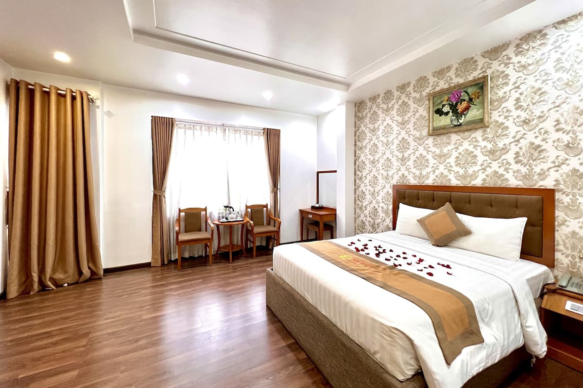 Une chambre d’hôtel avec un grand lit, un plancher en bois, un bureau avec des chaises, une fenêtre avec des rideaux et un mur orné d’un motif floral.