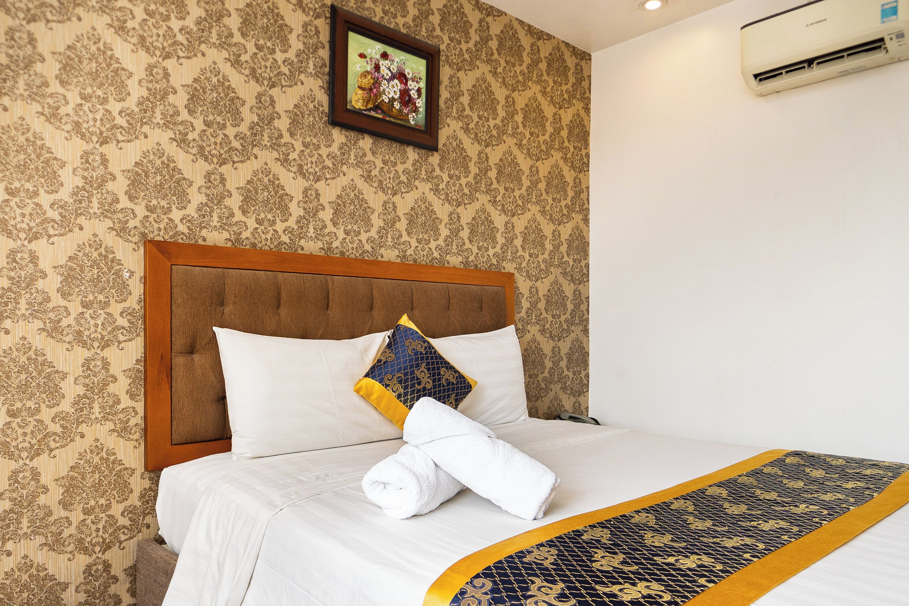 A hotel room with a bed, a headboard, a wall-mounted air conditioner, and a framed picture above the bed.