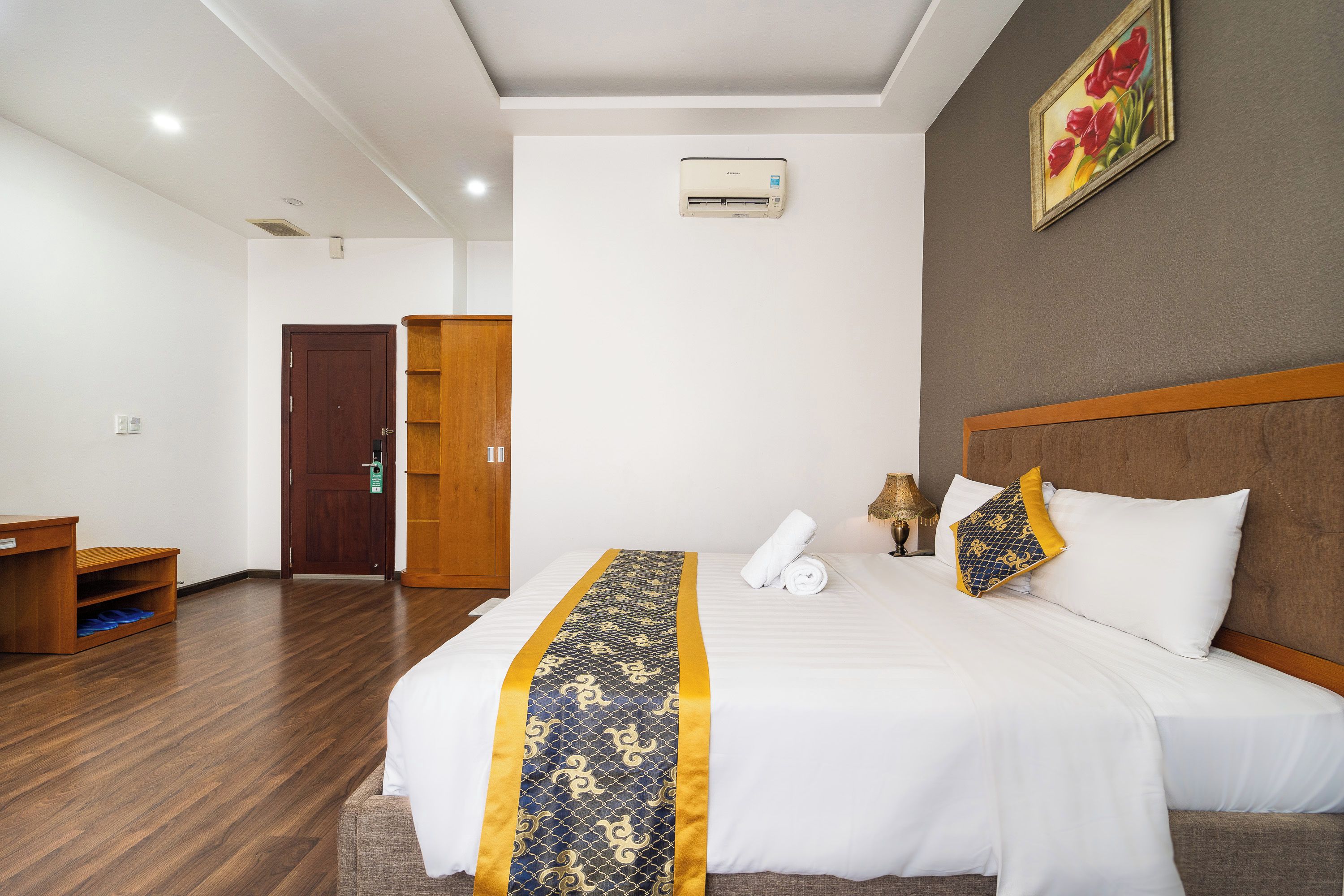 A hotel room with a bed, a wooden wardrobe, a desk, and a painting on the wall.