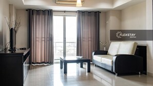 A modern living room with a sofa, a coffee table, and a desk. There is a large window with curtains, an air conditioning unit, and a sign for FlexStay Rentals. - Laidback Place Apartment (Bangkok)