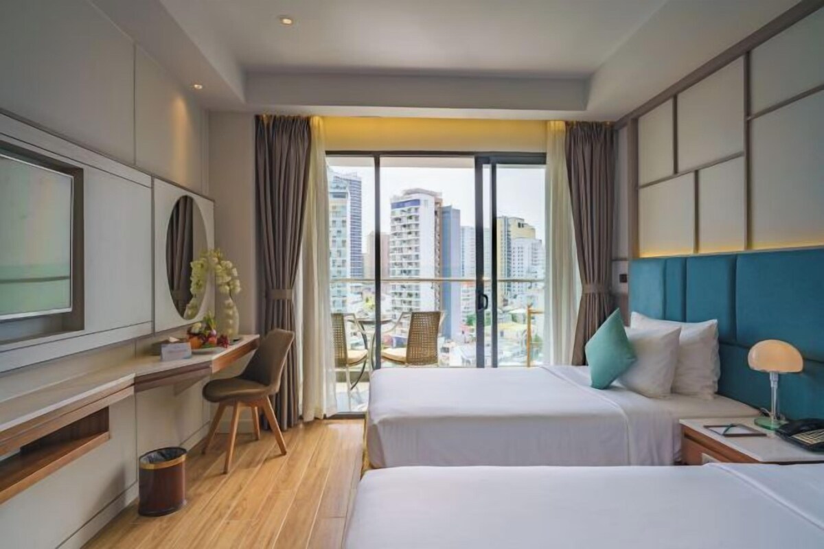 A hotel room with two beds, a desk, a TV, and a balcony with a city view.
