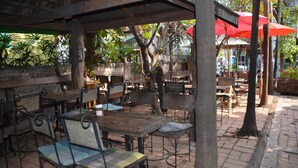 Un café al aire libre con mesas de madera y sillas de metal, rodeado de árboles y plantas en macetas.