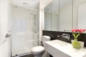 A modern bathroom with a glass shower enclosure, a white toilet, a white sink, and a mirror above the sink. - Manhattan Apartment (Melbourne)