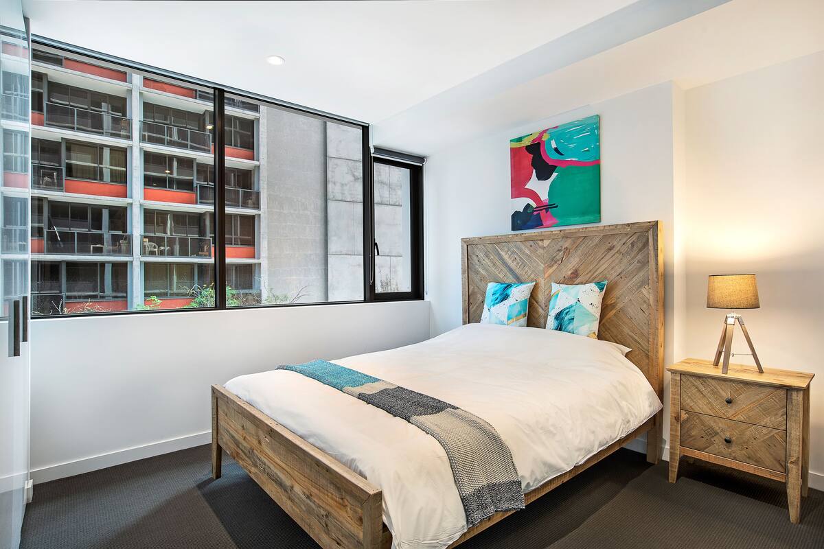 A modern bedroom with a large window, a wooden bed frame, a nightstand, and a colorful abstract painting on the wall.