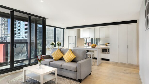 A modern apartment with a grey sofa, white coffee table, and a kitchen area with white cabinets and a microwave.