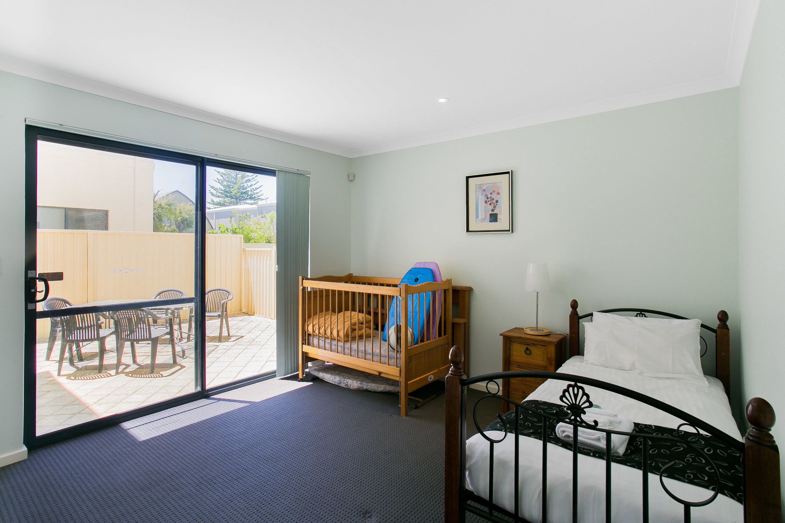 A room with a wooden crib, a bed with a black metal frame, a small wooden nightstand, and a large sliding glass door leading to an outdoor patio with chairs and a table.