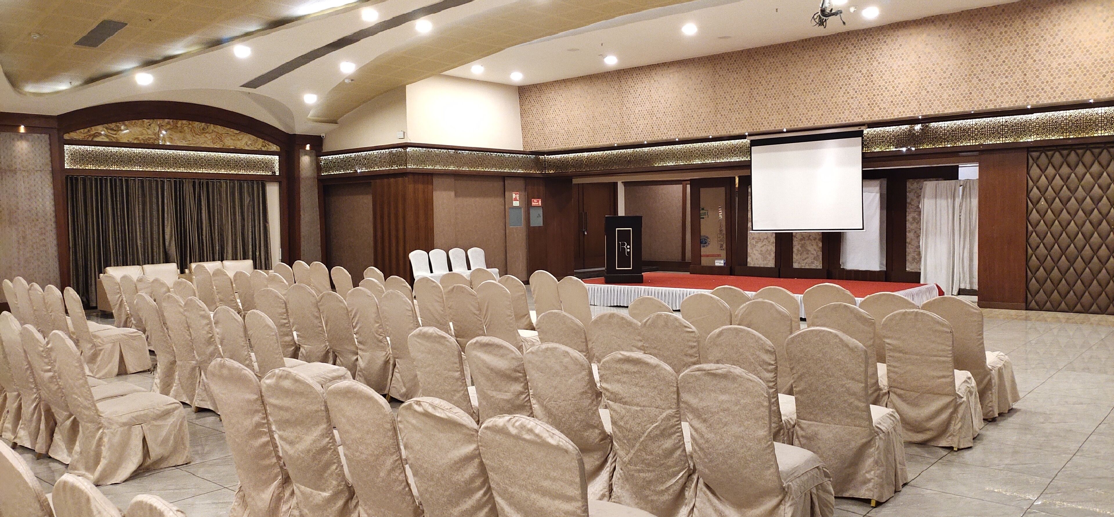 A conference room with rows of chairs covered in beige fabric, a podium, a projection screen, and a red tablecloth.