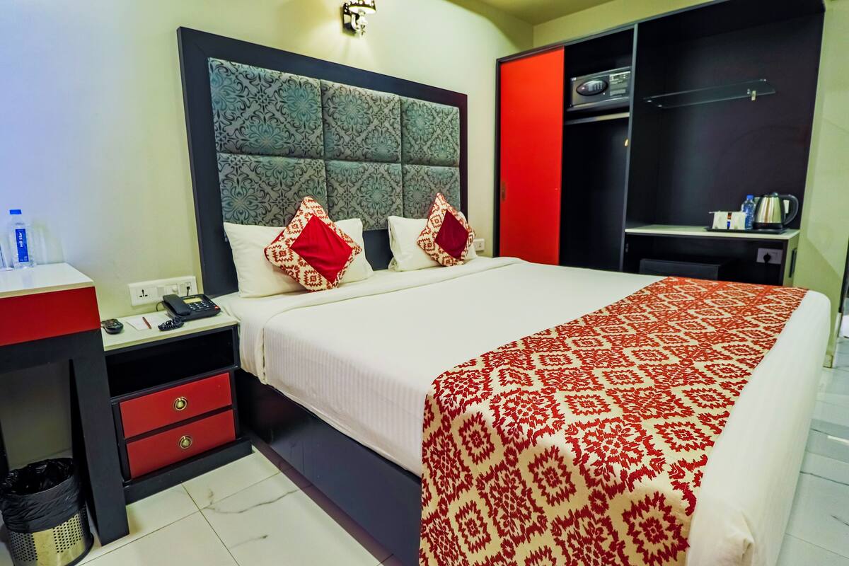 Habitación de hotel con una cama grande, una mesita de noche con un teléfono y un cubo de basura, un armario rojo y negro, y una colcha de rayas rojas y blancas.