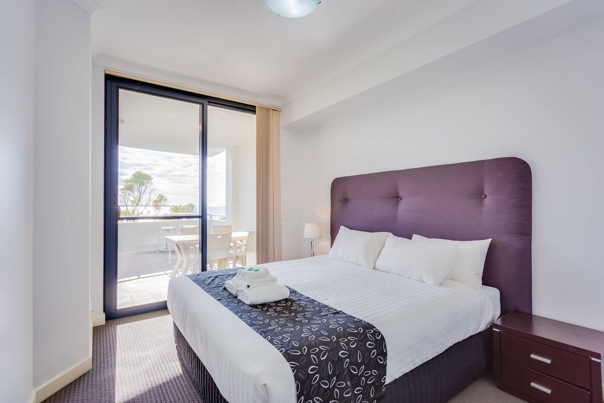 Ein Hotelzimmer mit einem großen Bett, einem Nachttisch, einer Lampe und Blick ins Freie.
