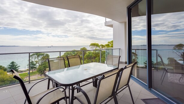 A balcony with a glass table and chairs, overlooking a body of water and greenery. - Nautilus by Rockingham Apartments (Rockingham)
