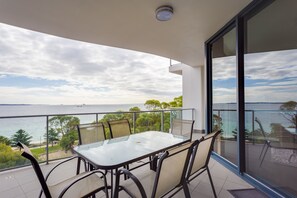 A balcony with a glass table and chairs, overlooking a body of water and greenery.
