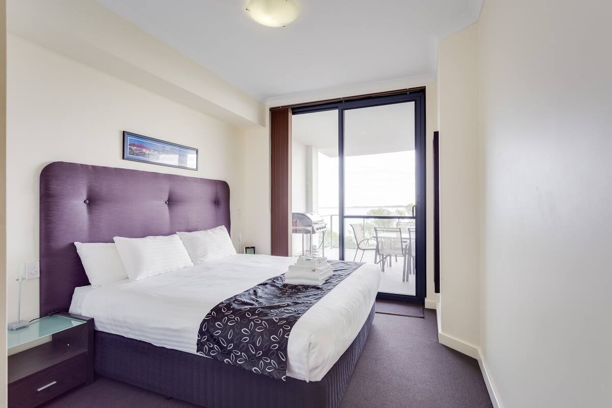 Ein Hotelzimmer mit einem großen Bett, einem Nachttisch, einem Sessel, einem Fenster und Blick ins Freie.