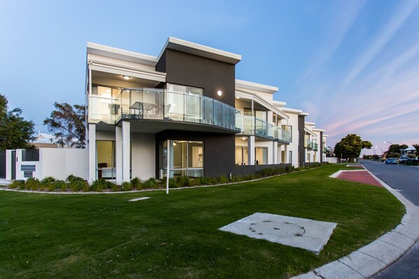 A modern multi-story residential building with balconies, a well-maintained lawn, and a clear sky.