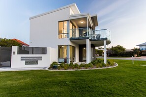 A modern two-story house with a balcony, a well-maintained lawn, and a gated entrance.