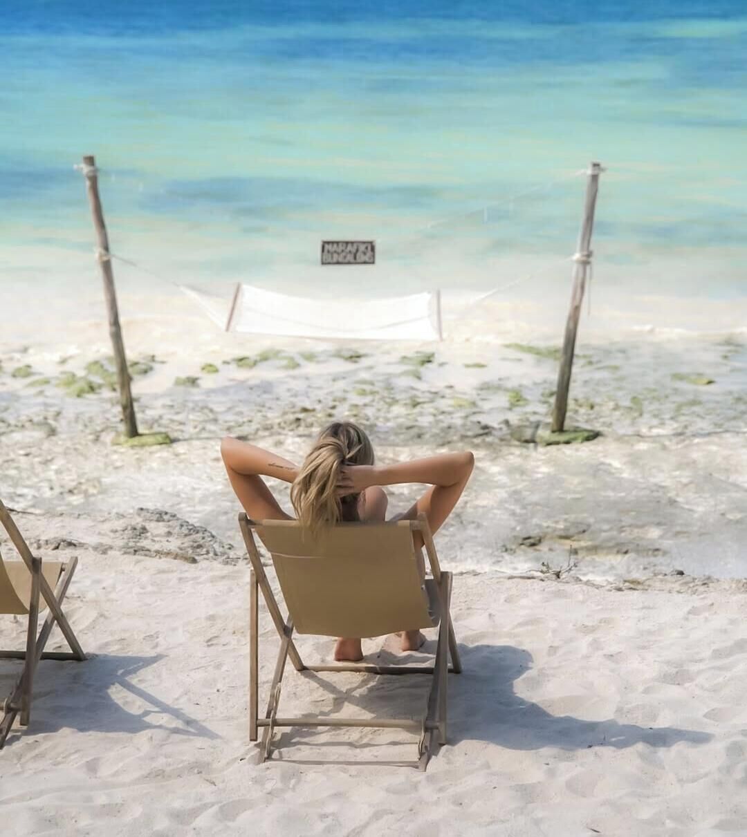 A beach scene with a person sitting on a chair facing the sea, a hammock, and a signboard.